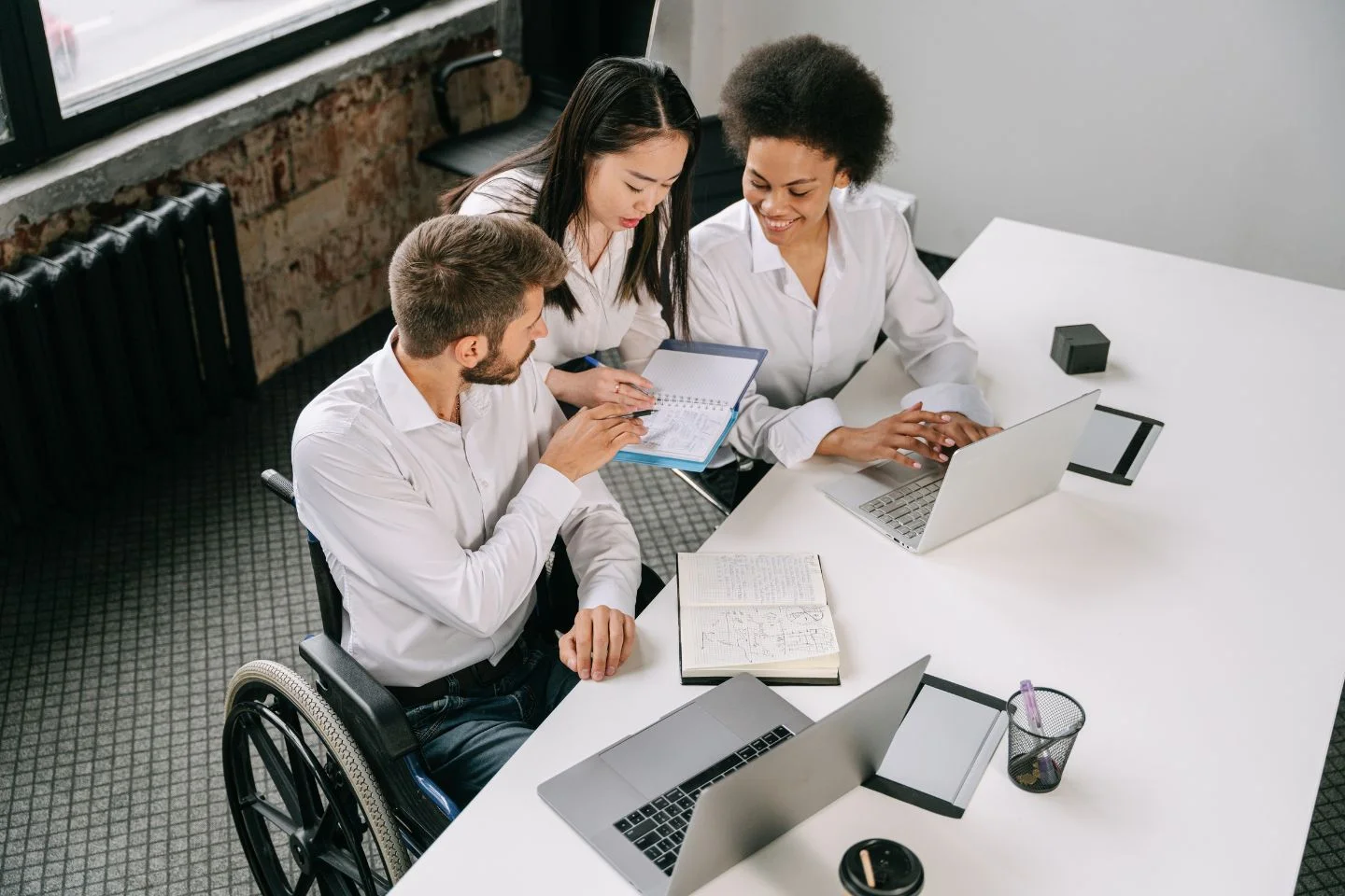 three people looking at a notebook, they are seated at the corner of a conference table with laptops in front of them