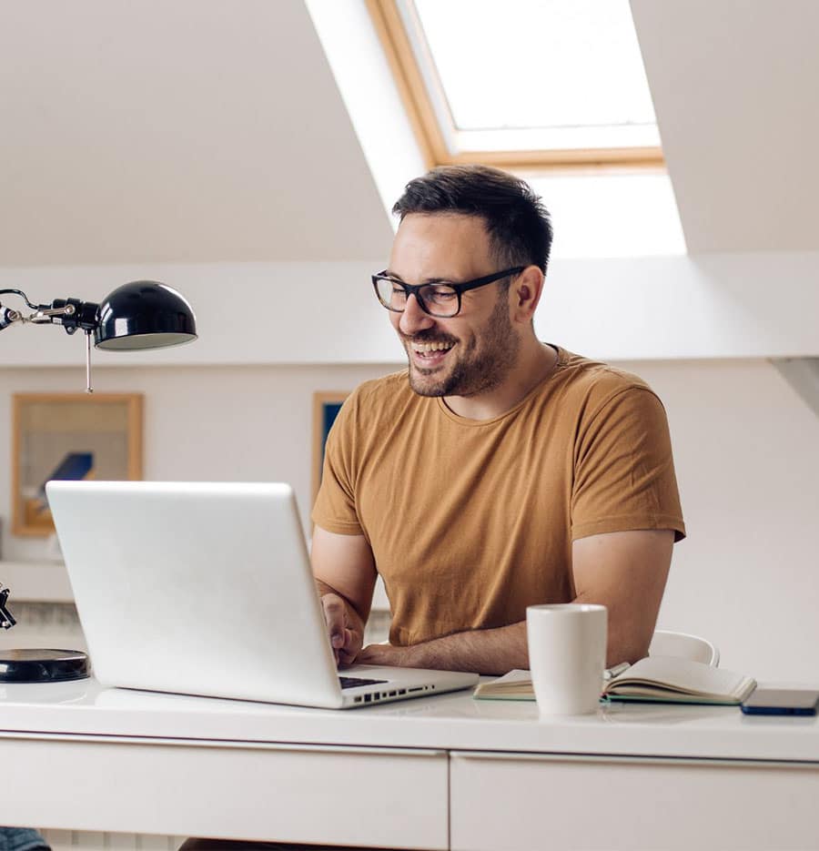 man with short brown hair, glasses, and a little bit of scruff sits at his home office desk, in front of a laptop; he's smiling at the screen