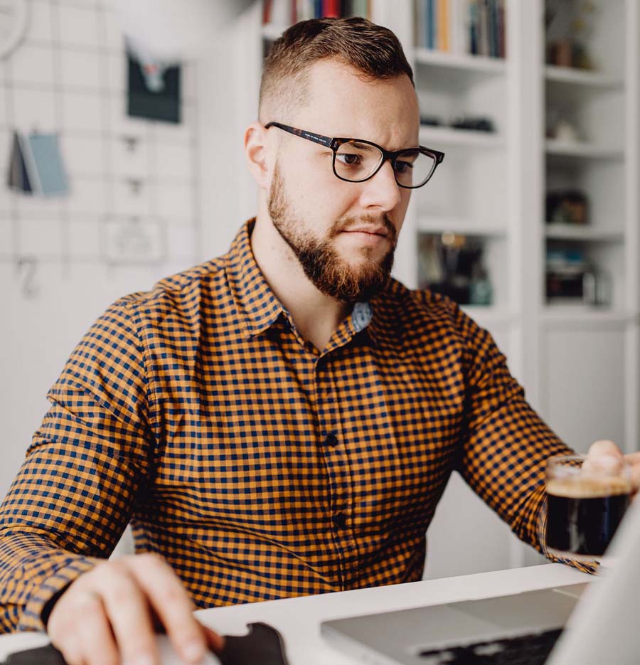 a man sitting at his desk in front of his laptop, he has one hand on a mouse while his other holds a cup of coffee, he is dressed casually