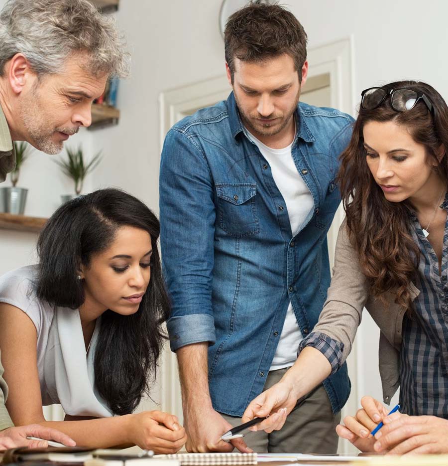 four individuals crowded around the corner of a table, looking at a paper to which the person on the right is pointing