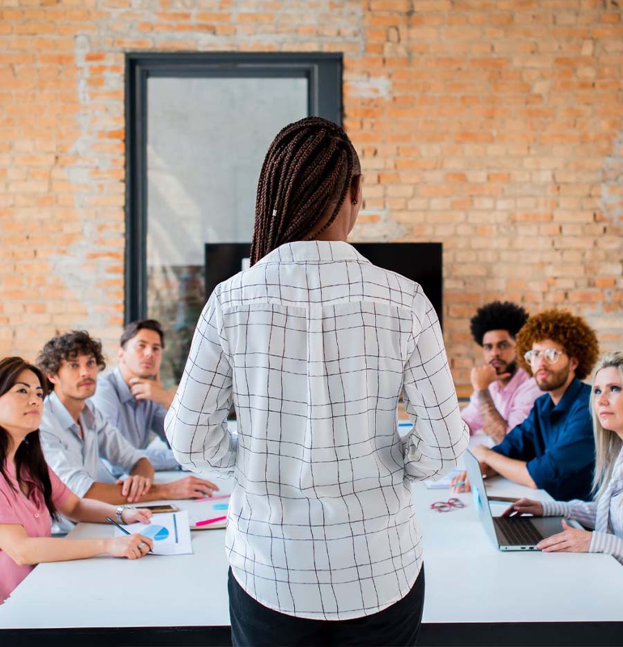 a person standing at the head of the table, we see them from behind while six diverse individuals at the conference table look at them